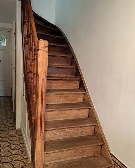 Escalier en bois avec rampe, mur peint en teinte claire et sol carrelé géométrique.