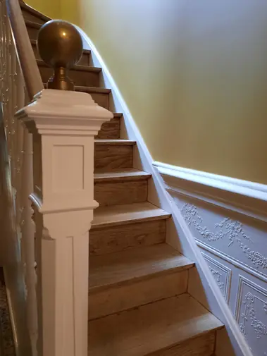 Escalier avec marches en bois, balustre peint blanc et boiseries moulurées sur mur jaune.