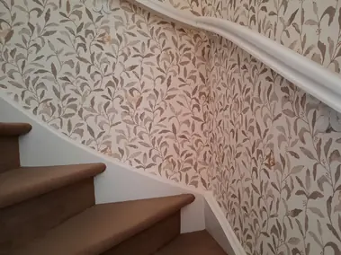 Papier peint motif feuilles et papillons sur escalier avec moquette brune et rampe blanche.