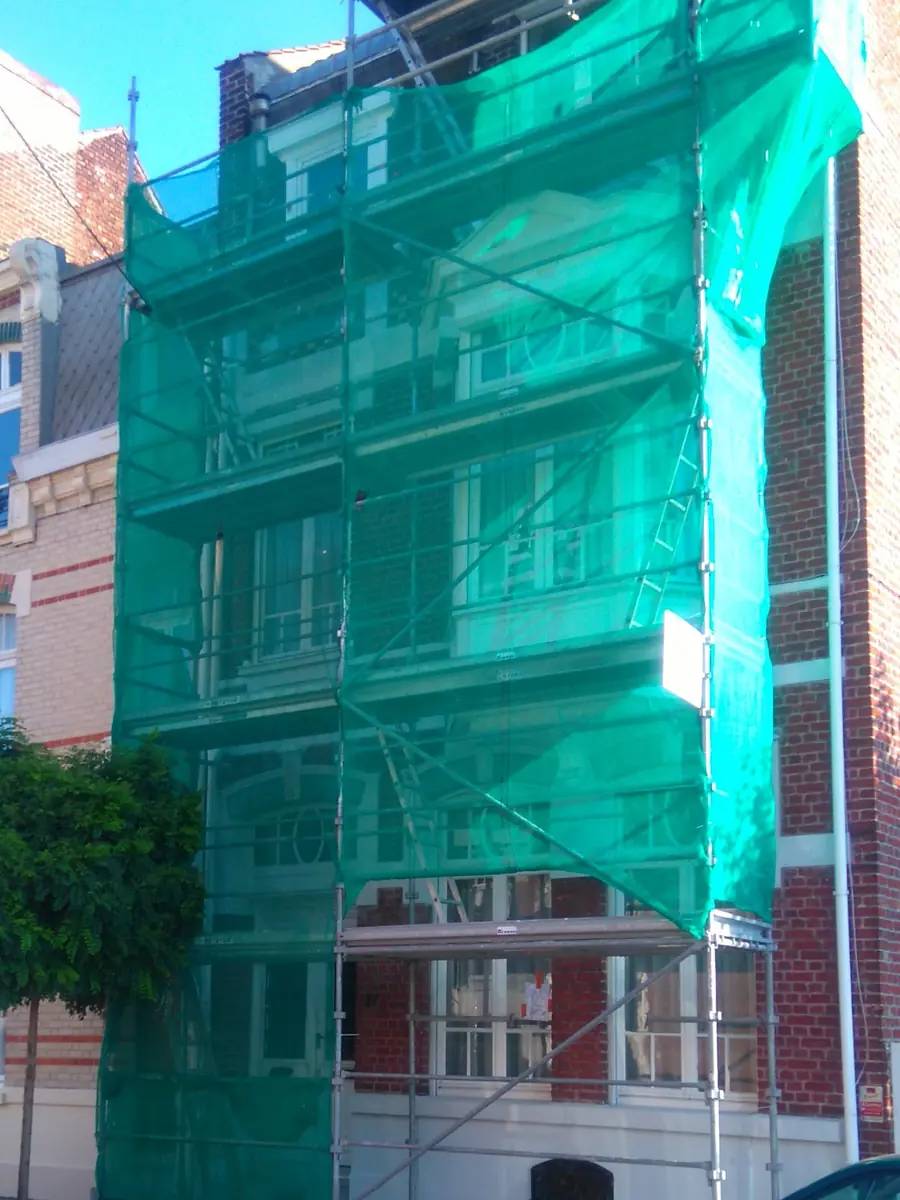 Façade en brique sous échafaudage avec filet de protection vert pour travaux de peinture extérieure.