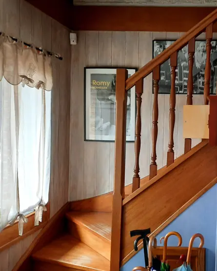 Escalier en bois avec balustres, murs lambrissés clairs, voilage blanc et peinture bleue sous l'escalier.